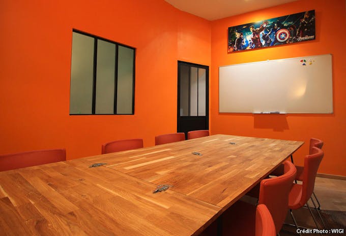 Une grande salle de réunion avec immense table en bois, de nombreuses chaises orange et tous les murs peints en orange. Sur le mur du fond, un tableau blanc.