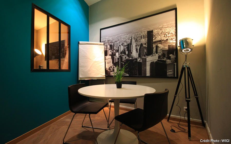 Une salle de réunion avec un mur bleu canard, une fenêtre verrière. Sur un mur, une immense photo en noir et blanc. Une table blanche avec 3 chaises et un paper-board dans le coin.