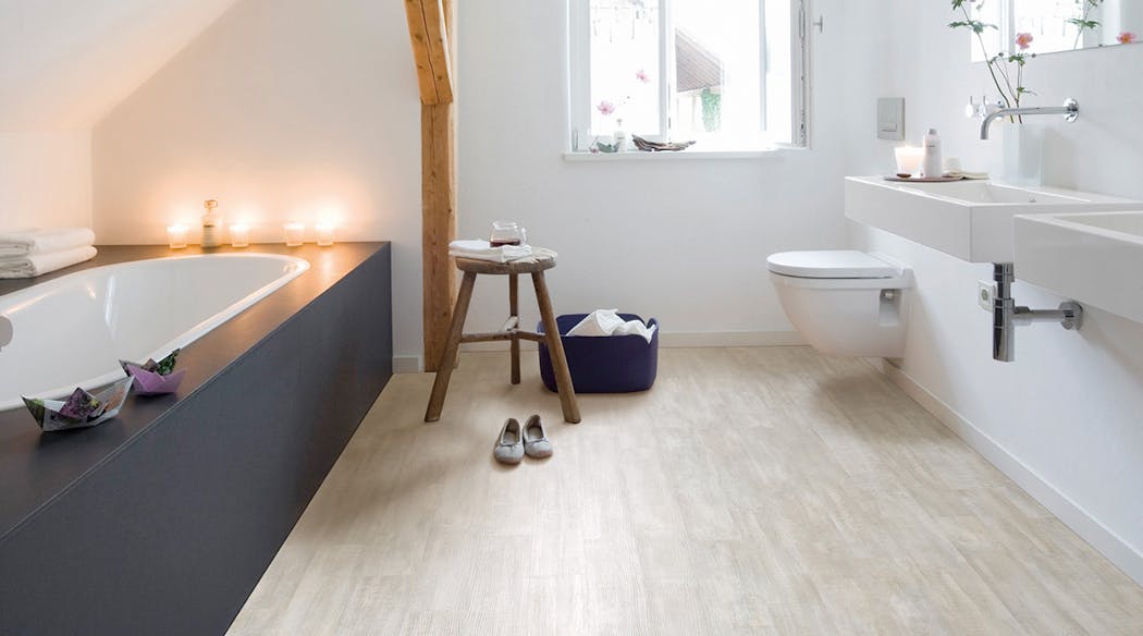 Une salle de bain sous pente avec une poutre. La baignoire est à gauche, un tabouret est posé sur un sol imitation bois très clair. Un lavabo est à droite.