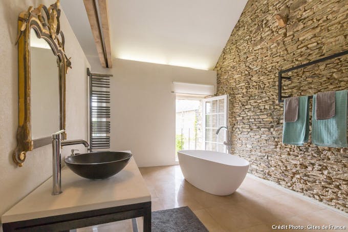 Une salle de bain très design avec une baignoire-îlot au milieu. A droite un mur de pierre apparente. A gauche, une vasque noire arrondie.