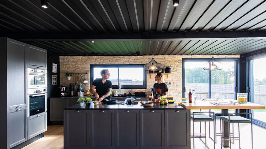 un couple prépare un repas dans une cuisine noire avec un ilot central et un plafond assorti.