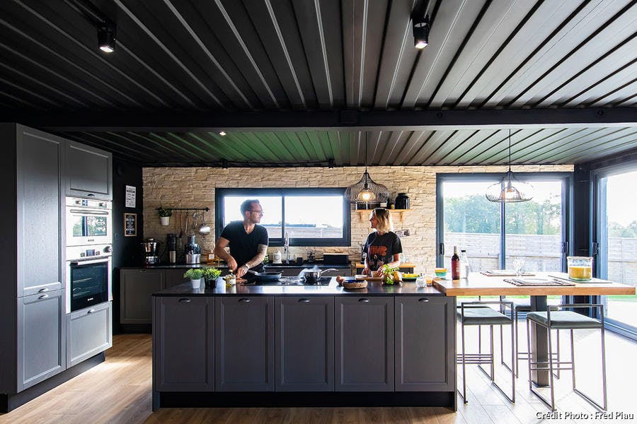 un couple prépare un repas dans une cuisine noire avec un ilot central et un plafond assorti.