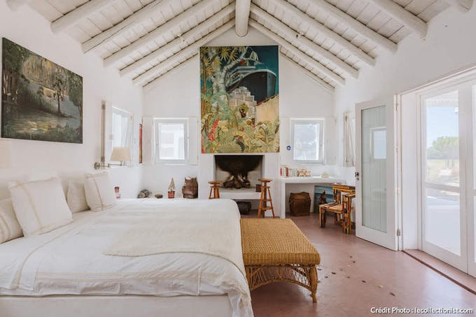 une chambre à coucher blanche avec une oeuvre d'art posée au-dessus de la cheminée et un banc en rotin