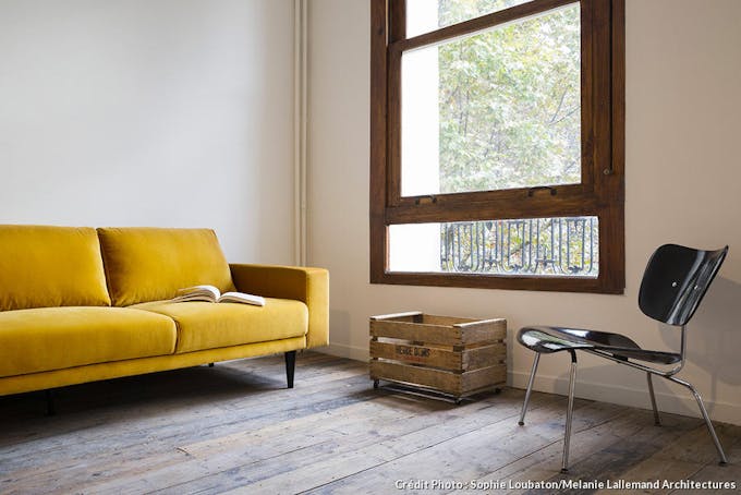 Dans un coin du salon, contre le mur de gauche, un canapé en velours jaune. Le mur de droite laisse apparaître une fenêtre à guillotine. A droite de la fenêtre, une chaise noire. Sous la fenêtre, une vieille caisse en bois à roulettes. Le sol est rec