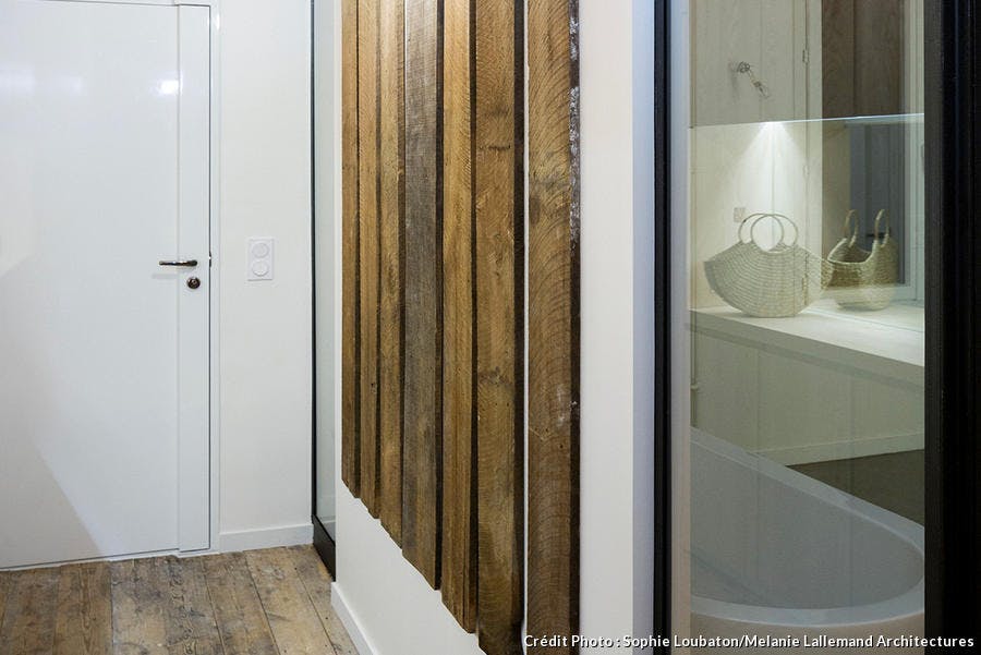 Dans l'entrée, des vieilles planches en bois fixées verticalement sur un mur blanc au milieu. À droite, on aperçoit la baignoire îlot de la salle de bain.