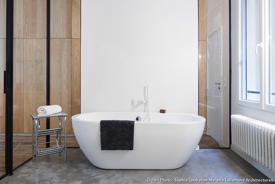 Dans l’espace salle de bains, au centre contre un mur, une baignoire îlot. À gauche de la baignoire, un porte-serviettes en métal argenté. Une serviette noire est posée sur le rebord de la baignoire. A droite, un vieux radiateur restauré et repeint e