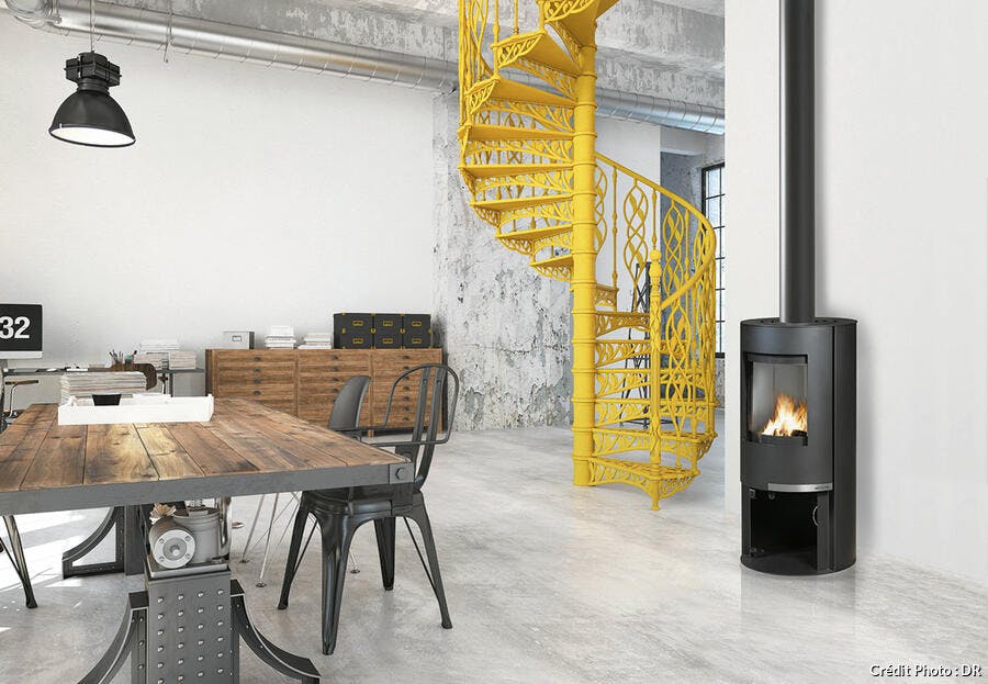une pièce à vivre de style industriel avec une grande table en bois et métal, des chaises en métal, un escalier jaune et un poêle contemporain