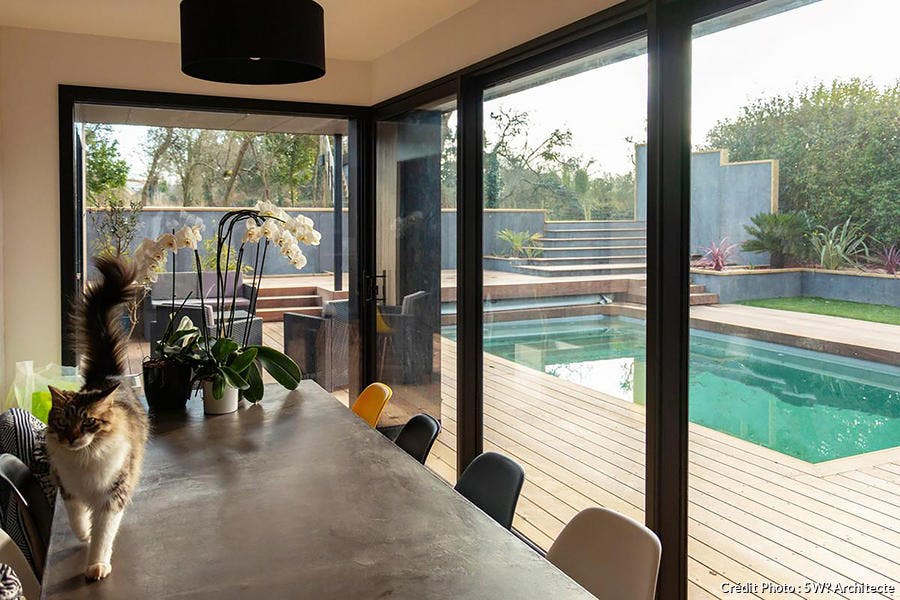 Une salle à manger avec des chaises Eames équipée de grandes baies vitrées donnant sur une piscine avec une terrasse en bois