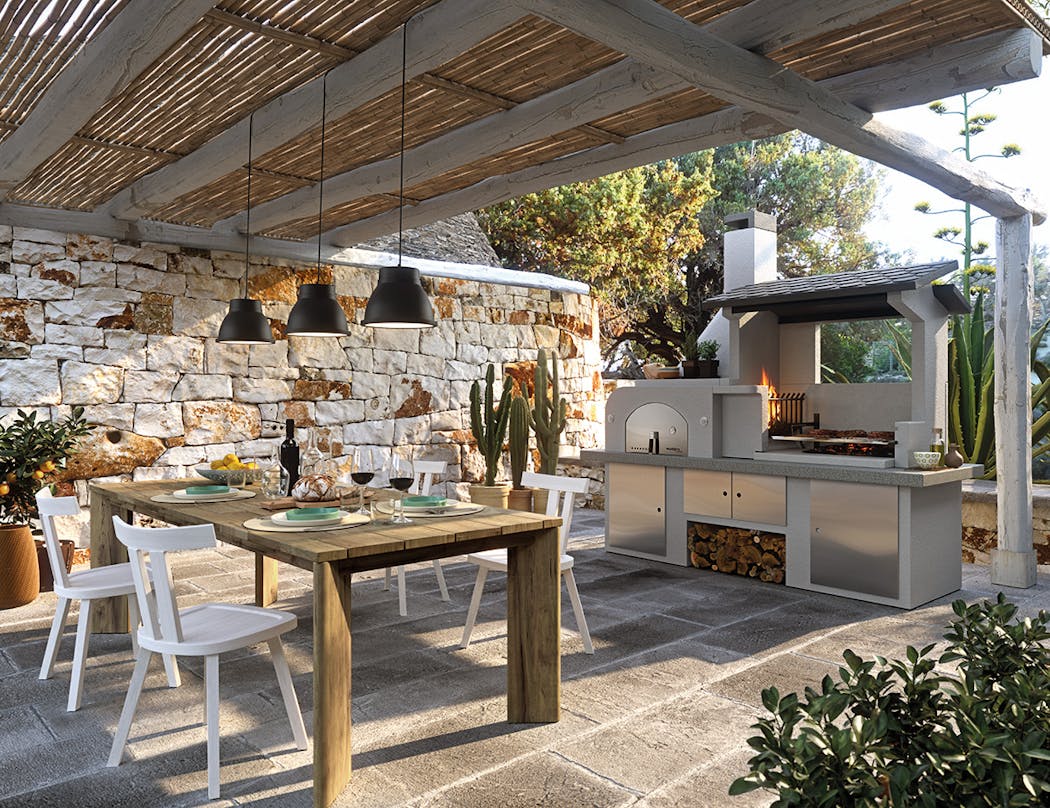 une salle à manger d'extérieur avec table en bois, suspension et cuisine au feu de bois sous une pergola