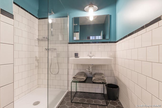 Une salle de bain avec vue sur la douche et le lavabo. Une partie des murs est en carreaux blancs, l'autre en peinture bleu canard. Au sol, des tomettes gris foncé.