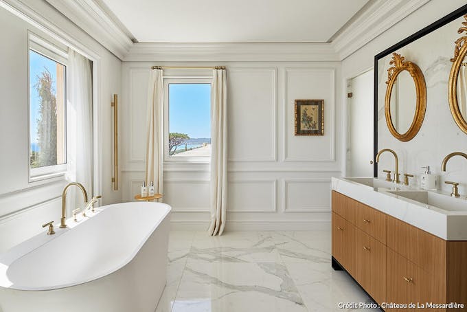 Une salle de bains blanche et bois avec sol en marbre et vue sur la mer.