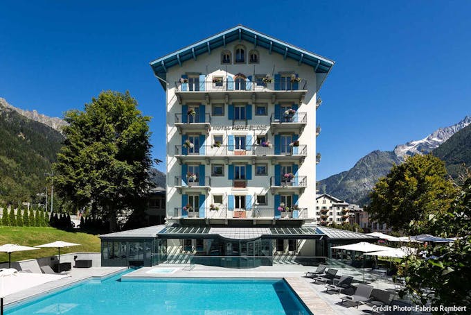 Vue d'ensemble de l'hôtel Mont-Blanc à Chamonix avec sa façade blanche, ses volets bleus sa piscine turquoise face aux montagnes.