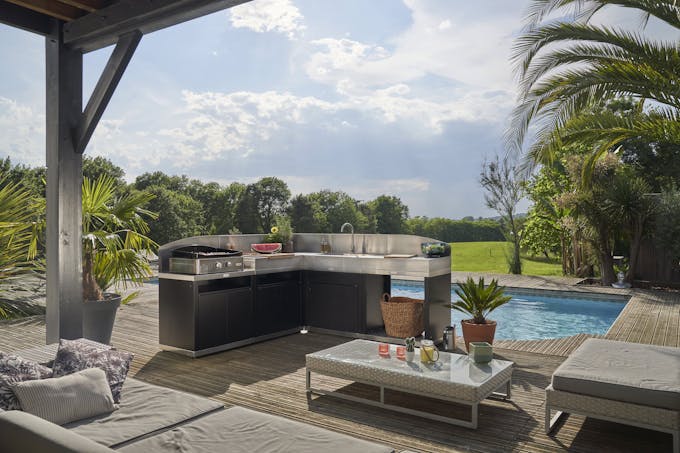 une cuisine extérieure en L équipée d'une plancha, installée dans un jardin devant une piscine.